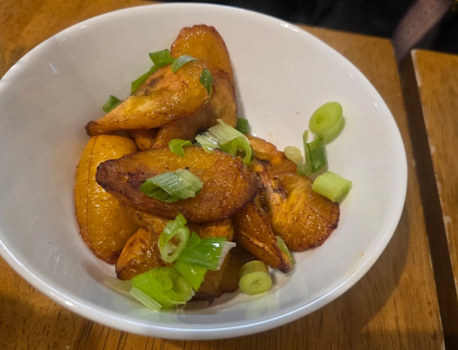 A white bowl of fried plantain wedges, caramelised dark at the cut edges, scattered with sliced spring onion