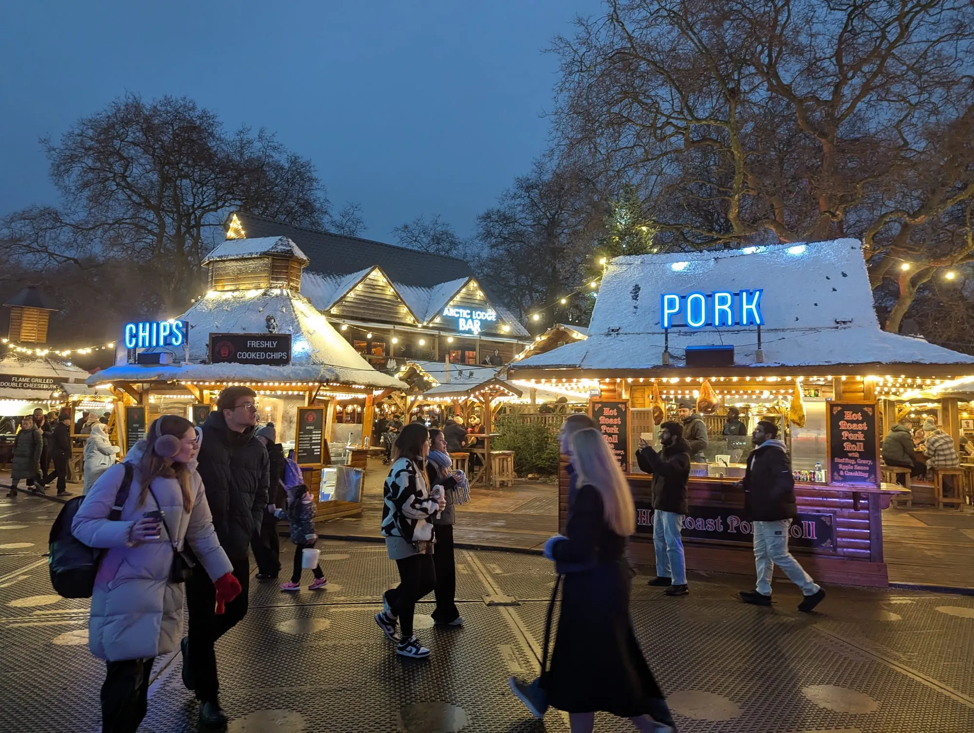 Food stalls lit up at dusk inside Winter Wonderland, selling chips and hot roast pork rolls, with a bar sign glowing behind