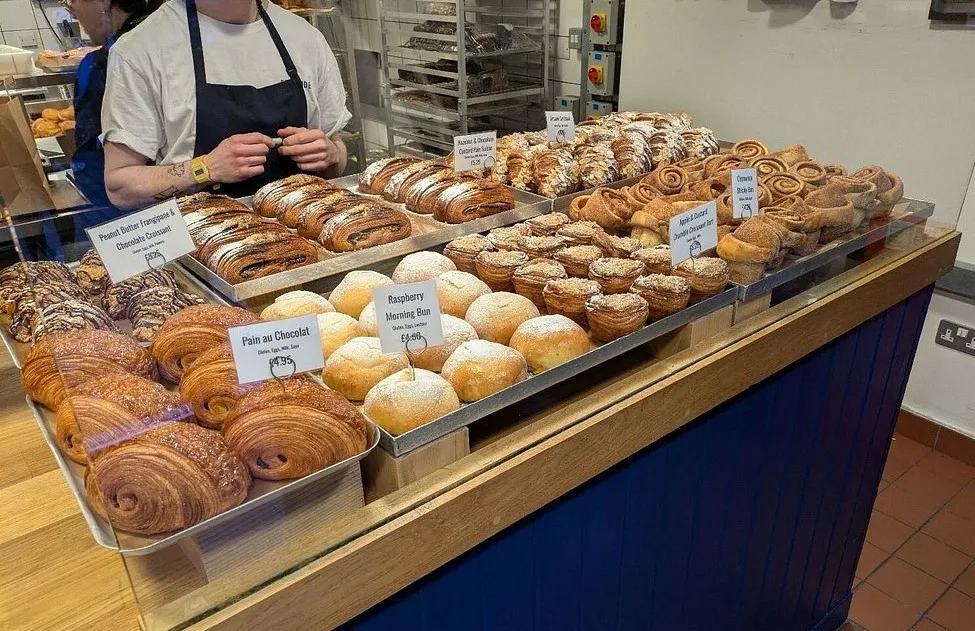Trays of pain au chocolat, sugar-dusted morning buns and cinnamon swirls on a bakery counter, with a baker in an apron working behind it