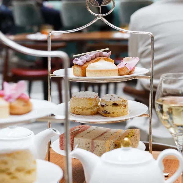 A tiered stand in the Fortnum's tea salon holding pastries, plain and fruit scones and finger sandwiches, with white teapots in front and green chairs behind