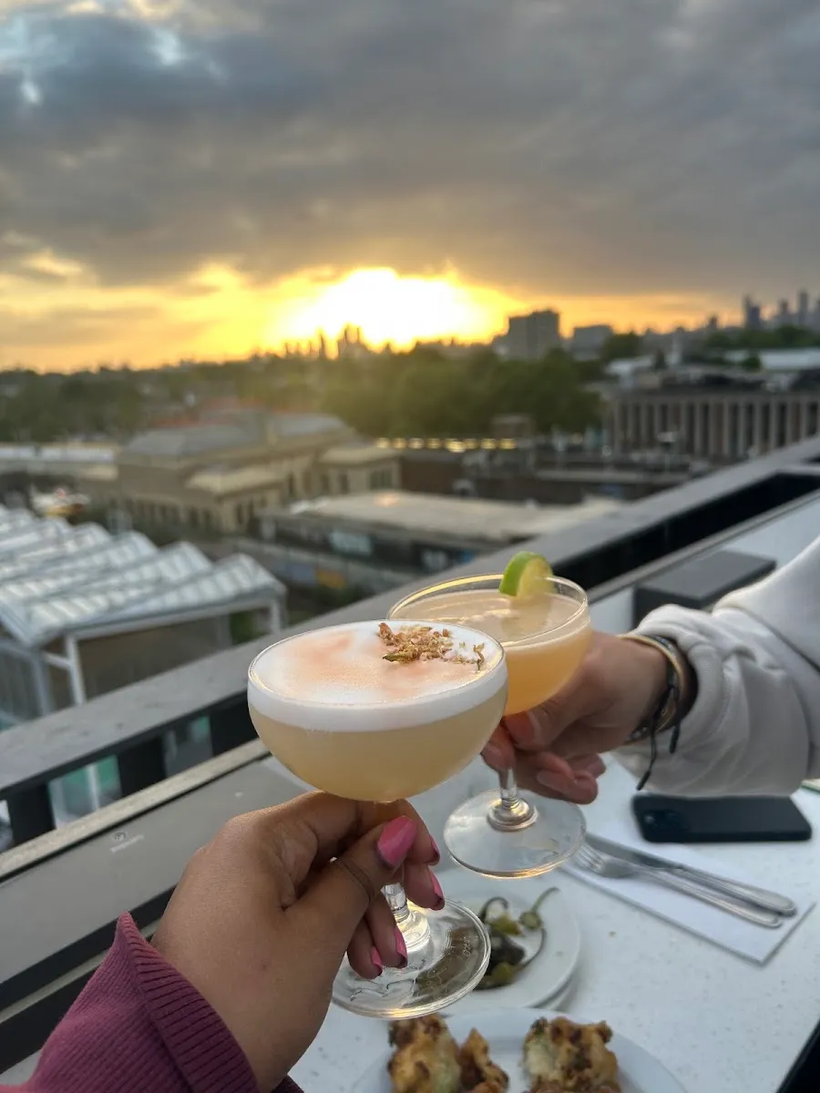 Two hands clinking cocktail glasses on a rooftop terrace at sunset, with the London skyline behind