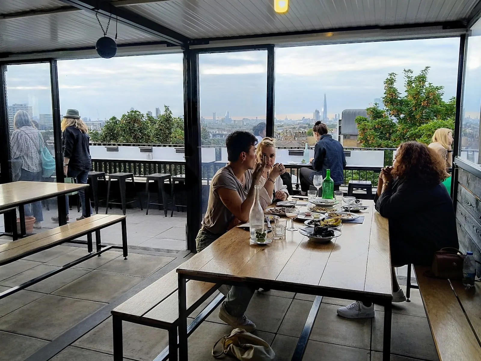 The top floor at Forza Wine in Peckham: long wooden refectory tables and benches under a low white ceiling, sliding glass onto an open terrace with bar stools, and the London skyline with the Shard on the horizon