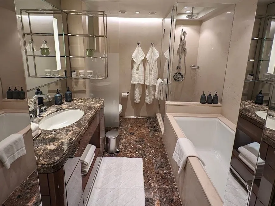 A Four Seasons Park Lane bathroom with a deep bath, a separate glass shower with a rainfall head, a brown marble vanity and two bathrobes on hooks