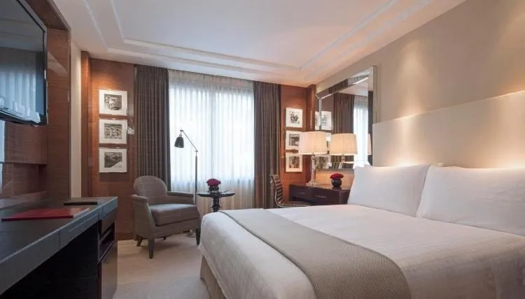 A Four Seasons Park Lane double room with a white upholstered headboard, a grey armchair and floor lamp by a tall curtained window, and framed black-and-white prints on a wood-panelled wall