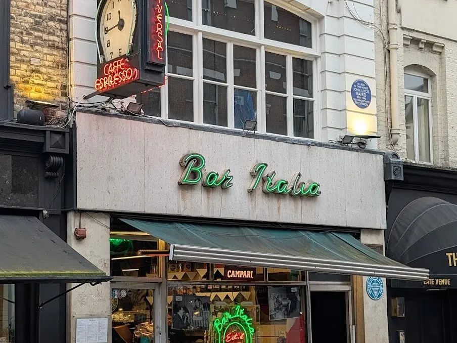 Bar Italia on Frith Street by day: green neon script on a stone fascia over a green awning, a hanging clock and Caffe Espresso neon above, and John Logie Baird's blue plaque on the wall to the right