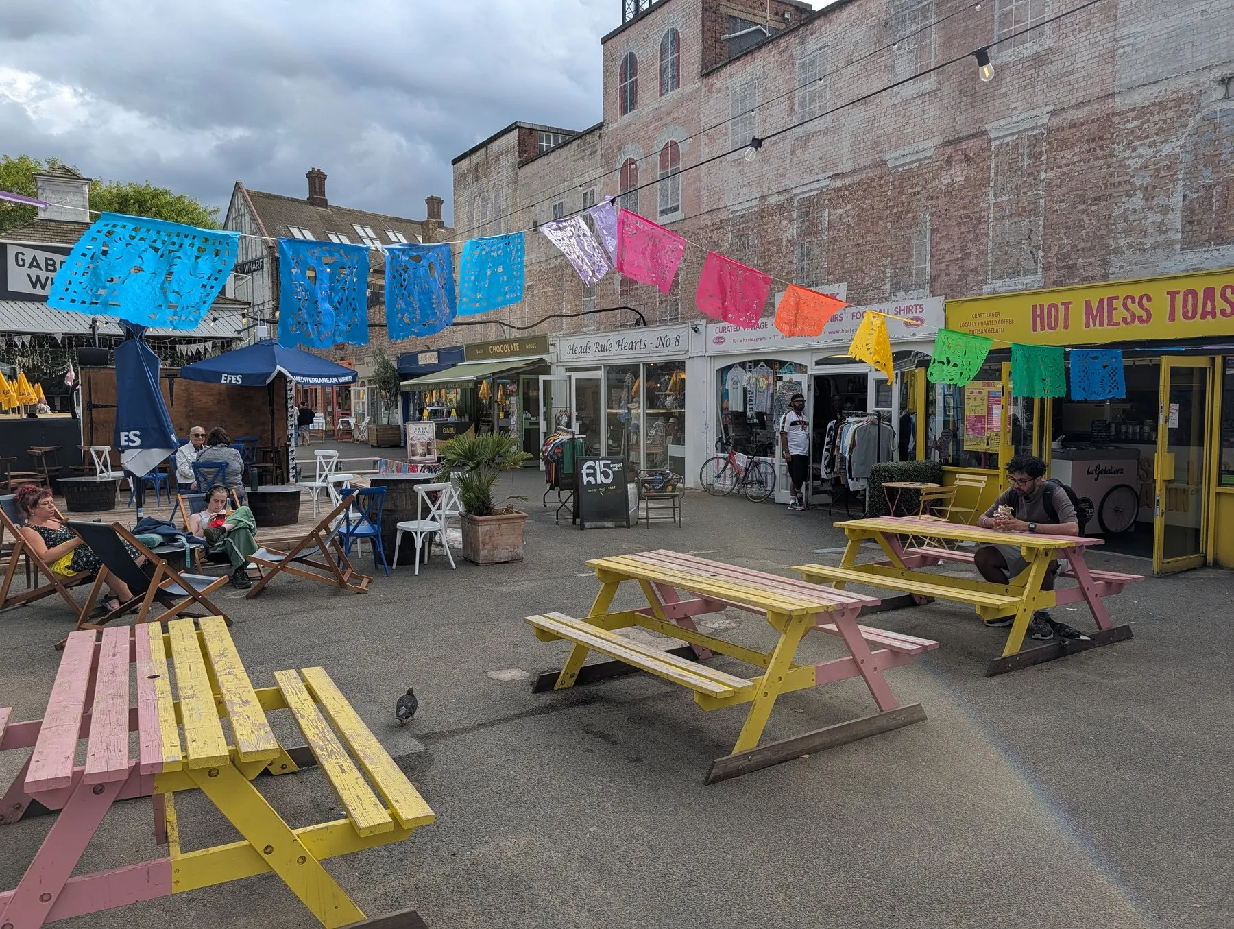 Painted picnic tables and deck chairs in the yard at Gabriel's Wharf, with papel picado bunting strung overhead and small independent units around the edge