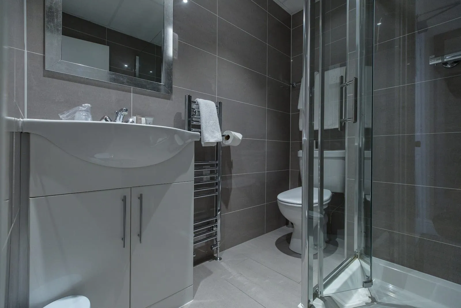 An en-suite at Garden Court Hotel with large grey tiles, a white vanity unit, a heated towel rail, a toilet and a corner shower cubicle with glass doors