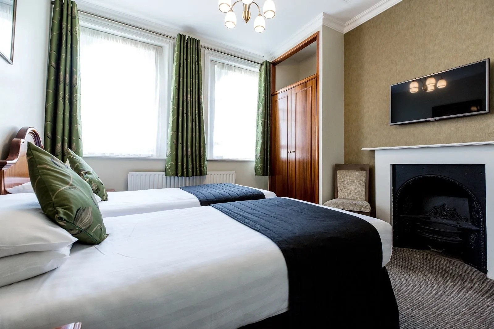 A twin room at Garden Court Hotel with two single beds under navy runners, green leaf-print curtains at two tall windows, a built-in wooden wardrobe and a black cast-iron Victorian fireplace