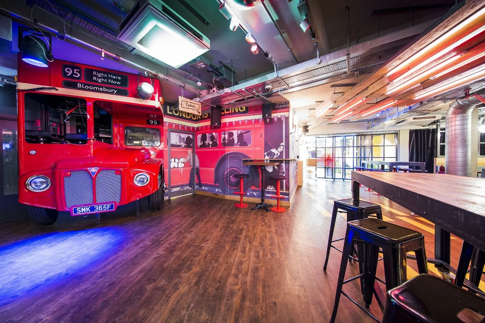 A red double-decker London bus converted into a bar inside the Generator London hostel lounge with seating and neon lighting