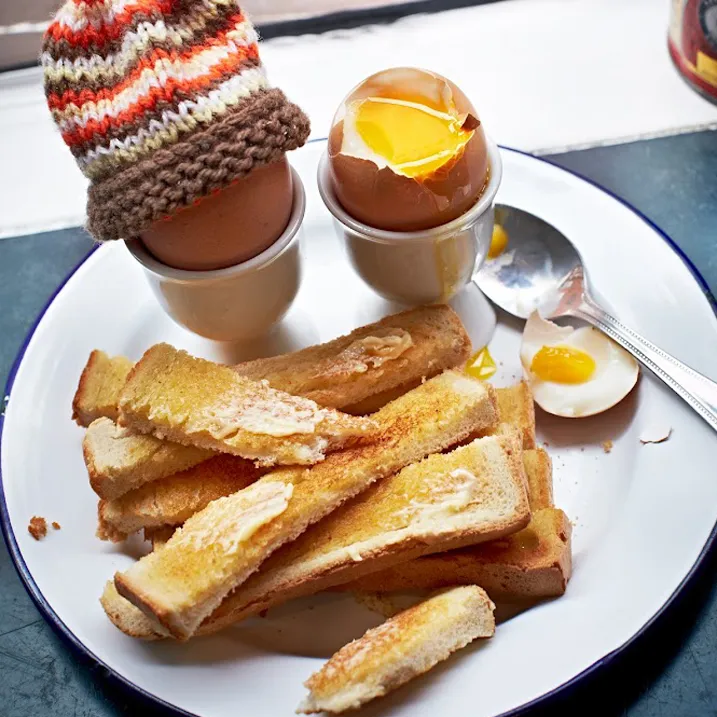 Two soft-boiled eggs in egg cups, one wearing a knitted cosy, with buttered toast soldiers at Ginger & White