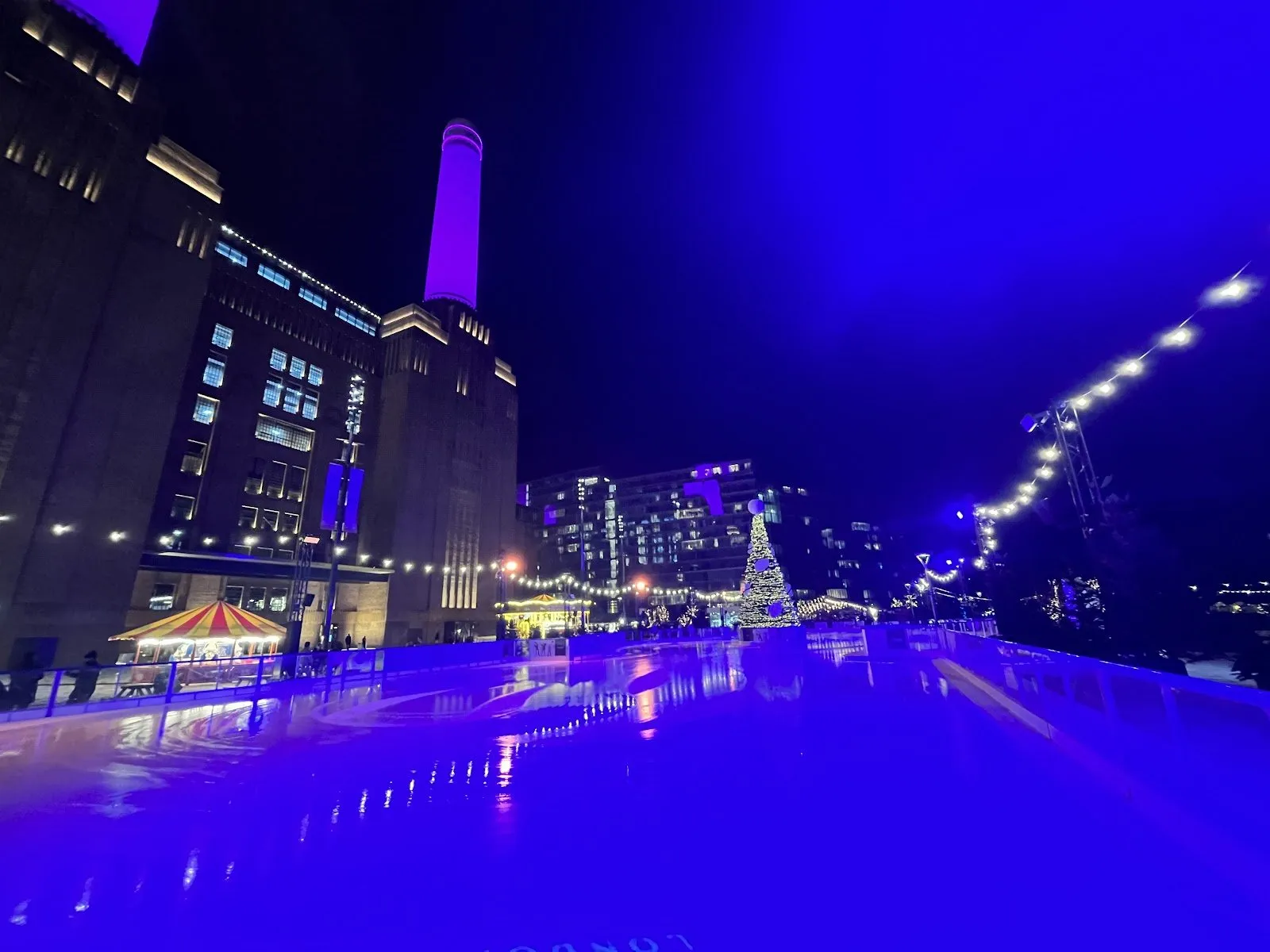 The Glide ice rink at night under blue lights, running alongside Battersea Power Station with a lit chimney, a striped carousel and a Christmas tree beyond
