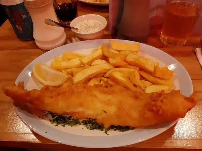 A long battered fillet and chips on a plate at Golden Union, with a lemon wedge, chopped parsley and a pot of tartare sauce on the table