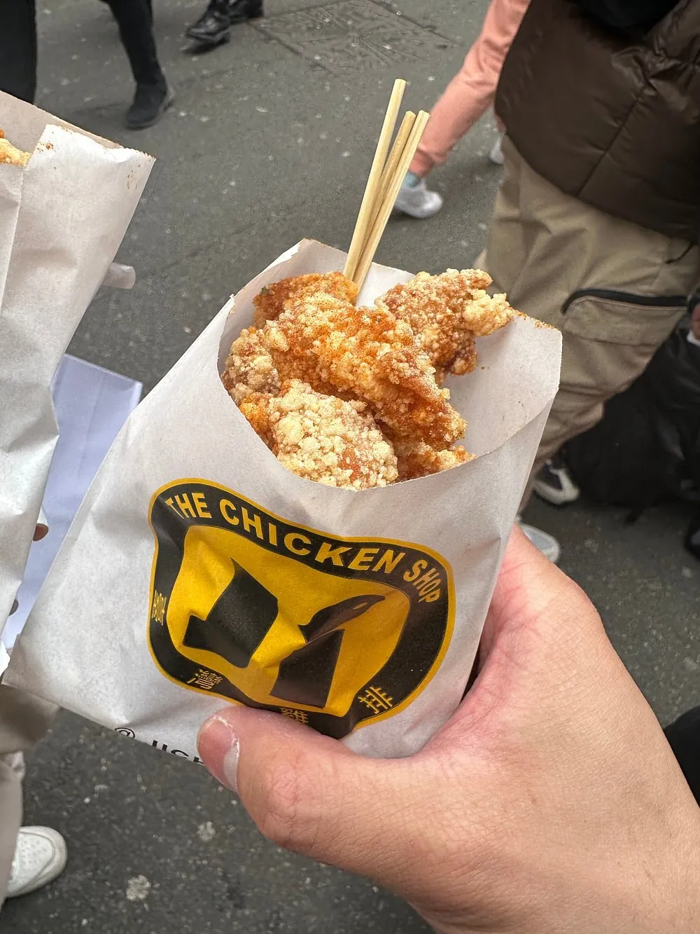 A paper bag of Taiwanese popcorn chicken held in one hand on a busy street, with wooden chopsticks pushed into it
