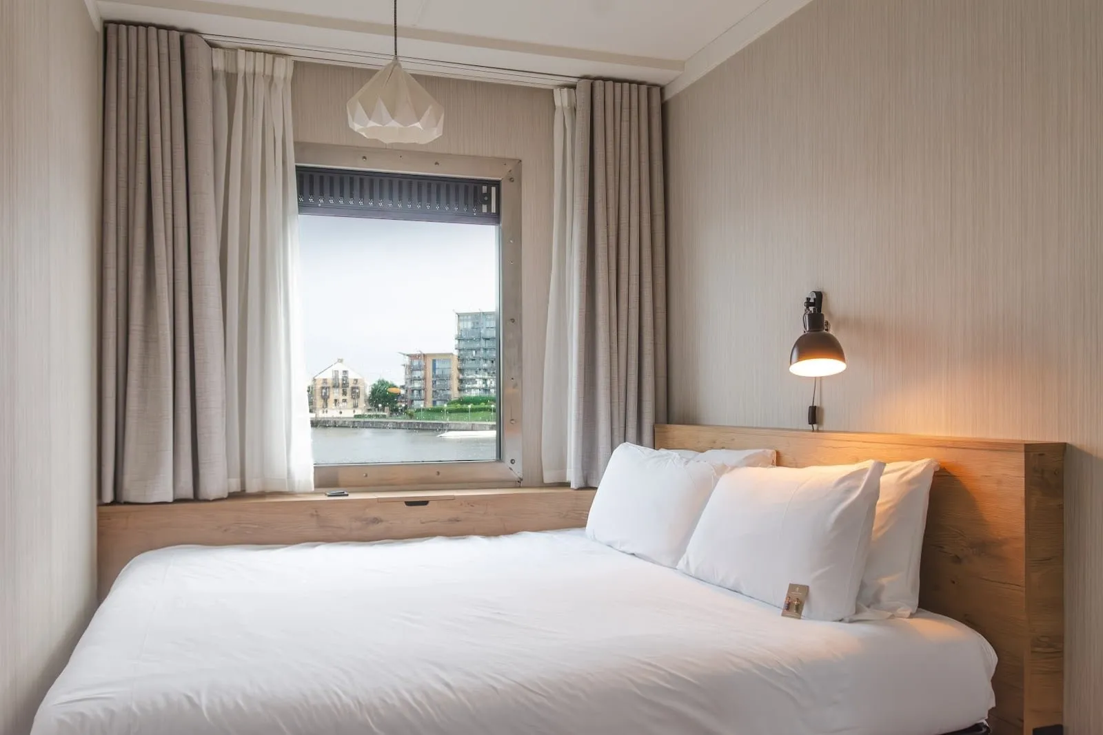 A double room at Good Hotel London with an oak headboard and a steel-framed window looking across Royal Victoria Dock