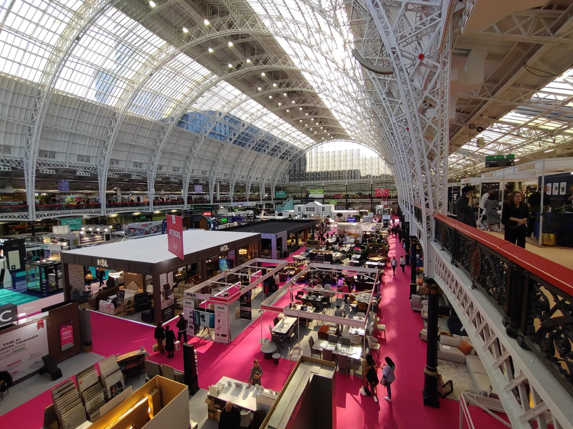 An aerial view across Olympia London's Grand Hall during the Ideal Home Show, exhibitor stands lining a pink-carpeted aisle beneath the hall's Victorian glass roof
