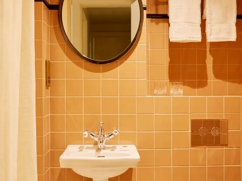 A bathroom at Grand Hotel Bellevue London with peach tiles, a small white basin with cross-head taps, a round mirror and a cream shower curtain