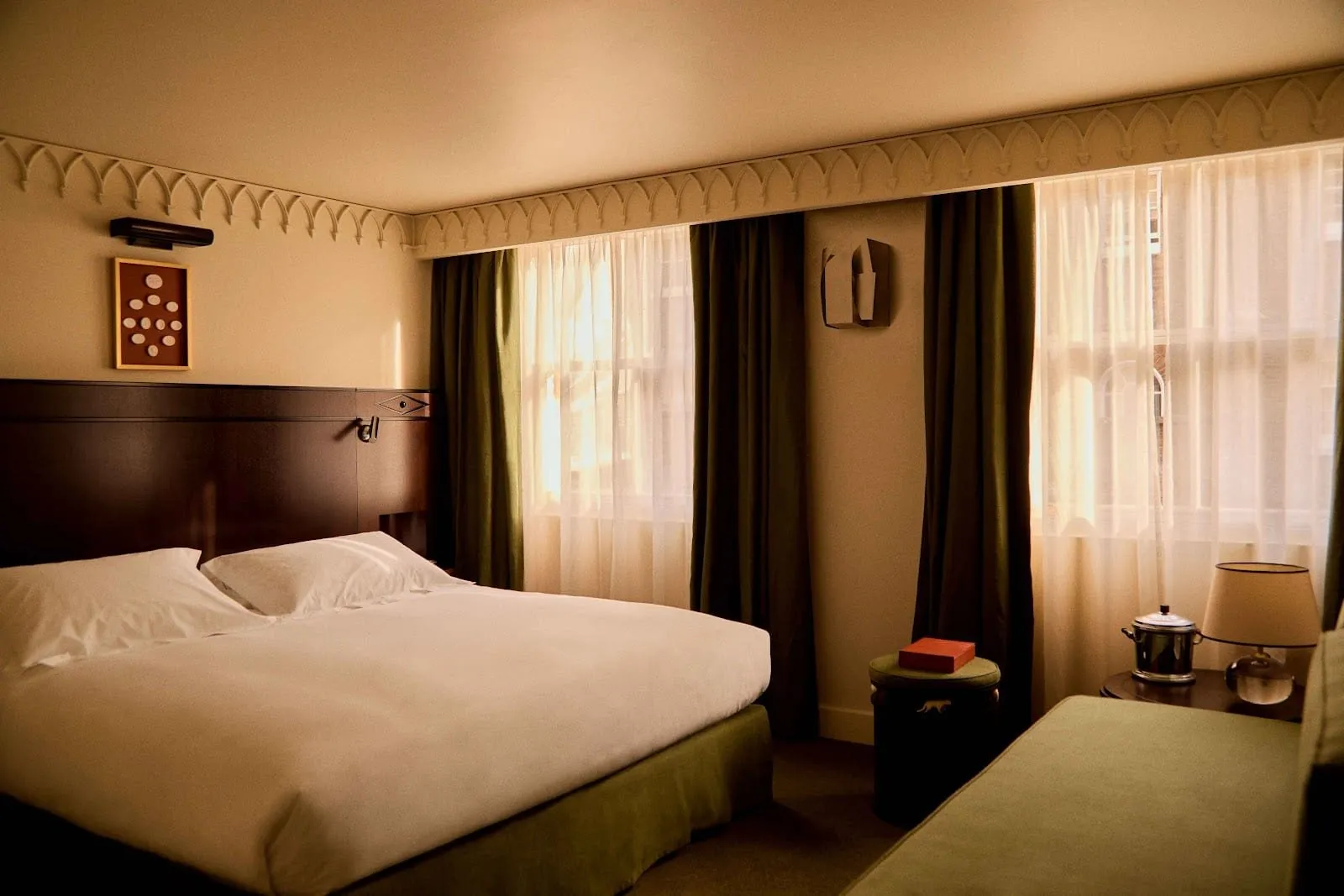 A double room at Grand Hotel Bellevue London with a dark wood headboard, two sash windows behind olive-green curtains, a Gothic-arch cornice and a green upholstered sofa