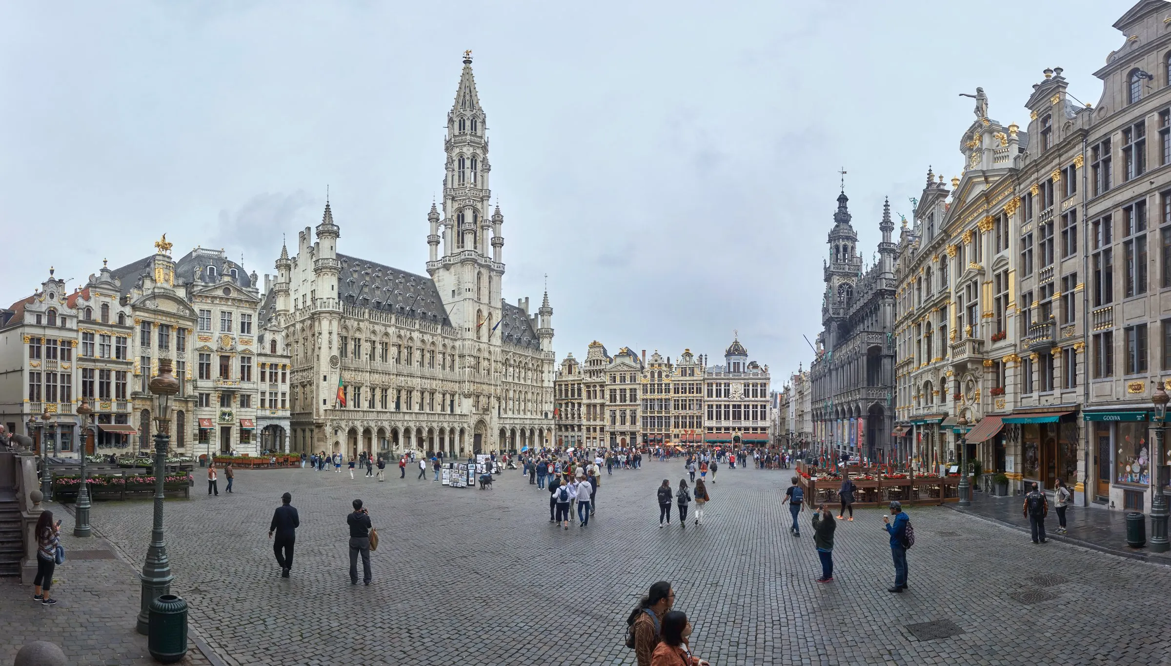 A wide panorama of the Grand-Place in Brussels, the Town Hall's spire rising above the gilded guildhalls as tourists cross the cobbled square
