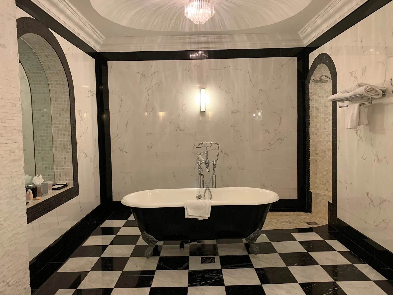 A suite bathroom at Grand Royale Hyde Park with a black claw-foot bath in the middle of a black and white chequerboard marble floor, white marble walls, a crystal chandelier and an arched walk-in shower