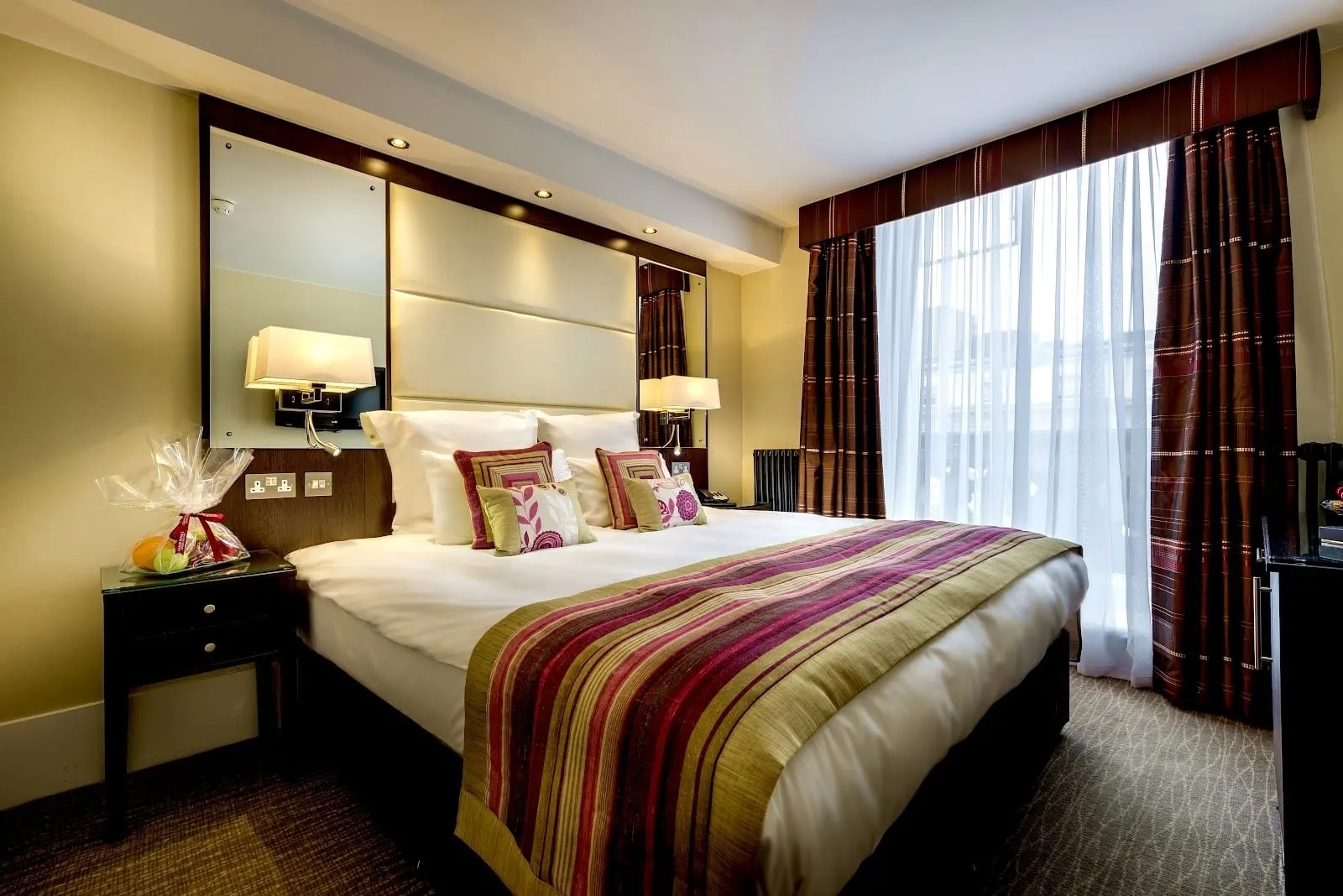 A double room at Grand Royale Hyde Park with a tall cream padded headboard between mirrored panels, a striped plum and olive bed runner, wall lamps and a full-height window with red striped curtains