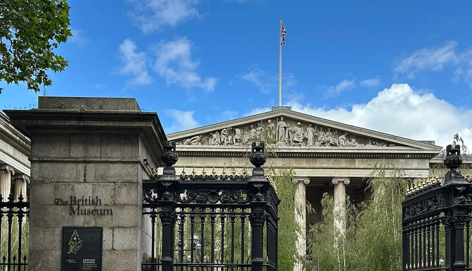 The black and gold railings and a stone gate pier carrying the museum's name, with the pediment and flagpole above the trees