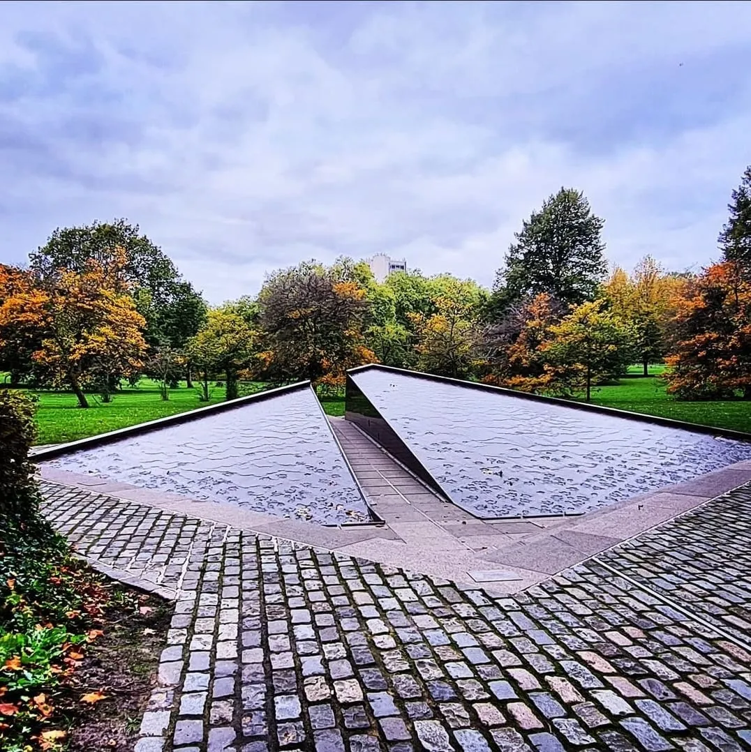 The Canada Memorial in Green Park, water running over sloped red granite, with trees turning gold and orange behind it
