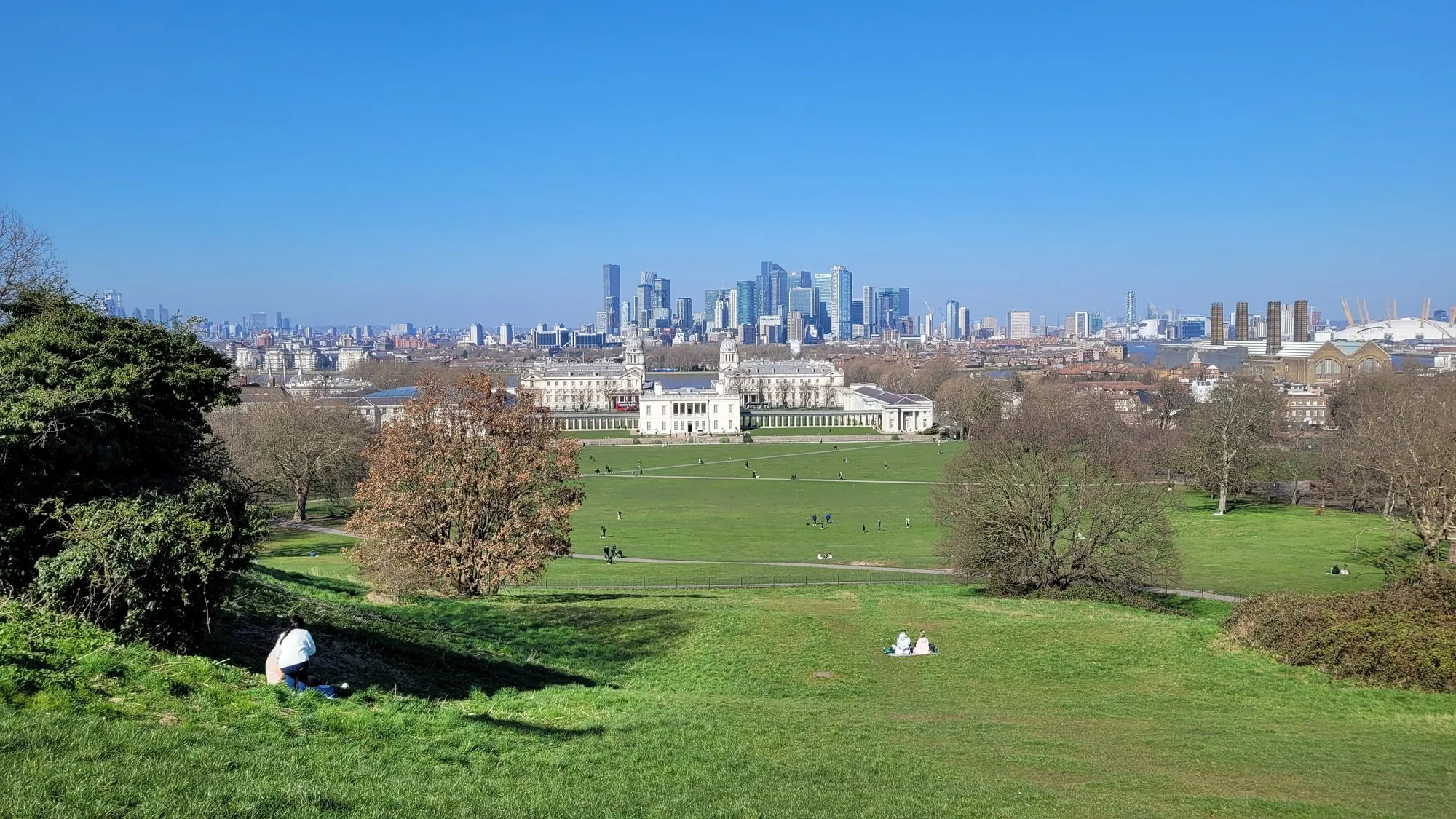 Greenwich Park in spring, looking down the grass slope over the Old Royal Naval College to Canary Wharf and the O2 under a clear blue sky