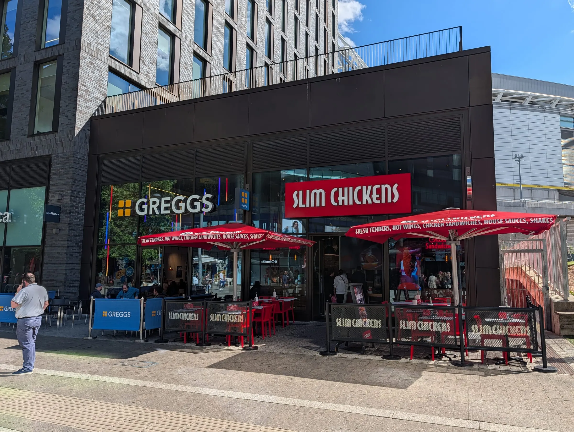 Greggs and Slim Chickens side by side on the ground floor of a modern block, with red umbrellas and outdoor tables in front