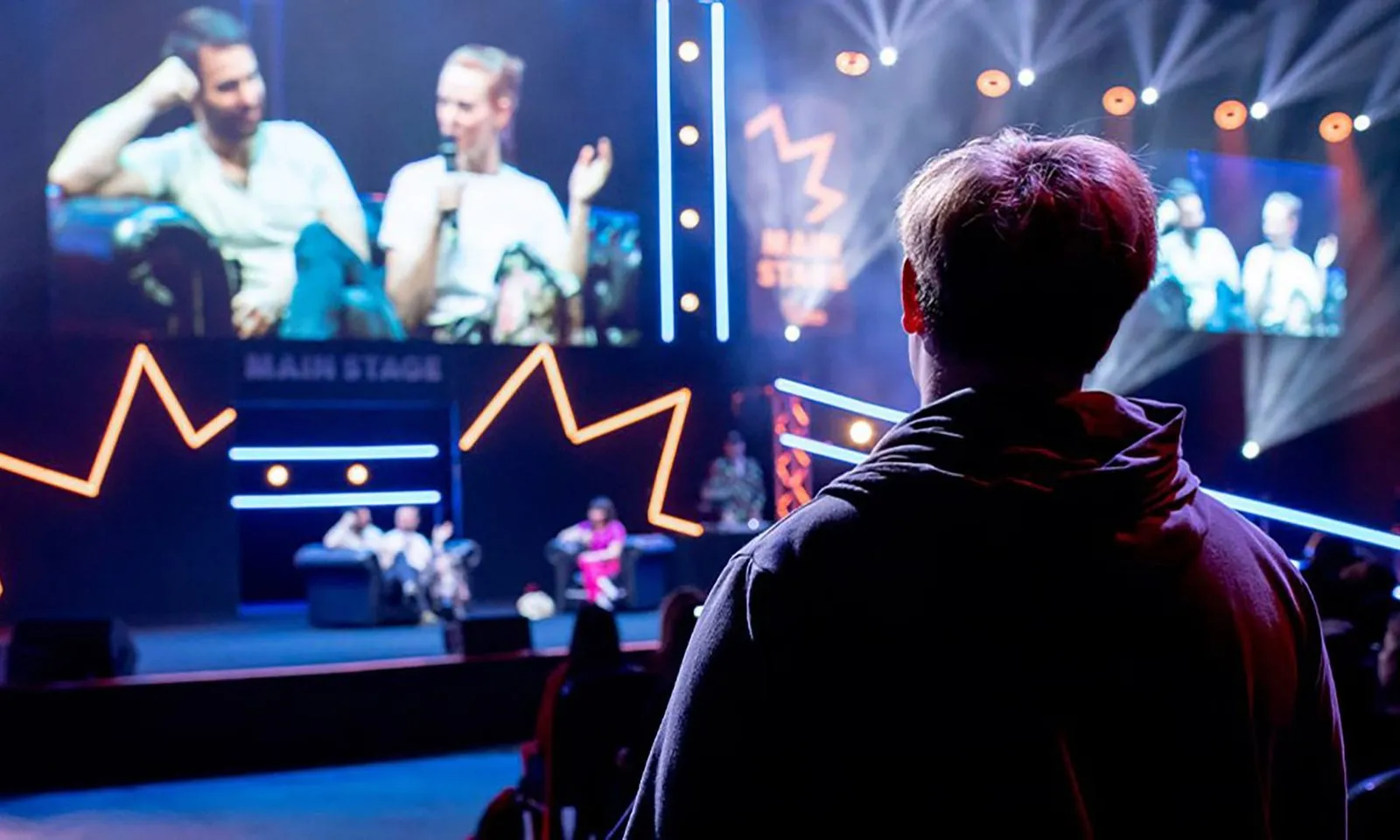 A fan watching a guest panel on MCM's Main Stage, with the panel showing on the big screens behind