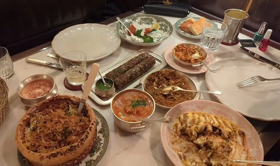 A full table at Gymkhana: a biryani sealed under a browned pastry lid, seekh kebabs with green chutney, a curry in a copper handi and a whole spiced cauliflower, on patterned china