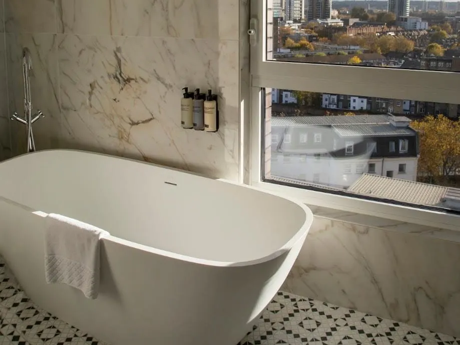 A freestanding bath beside a large window at H10 London Waterloo, with marble walls and a black-and-white patterned floor