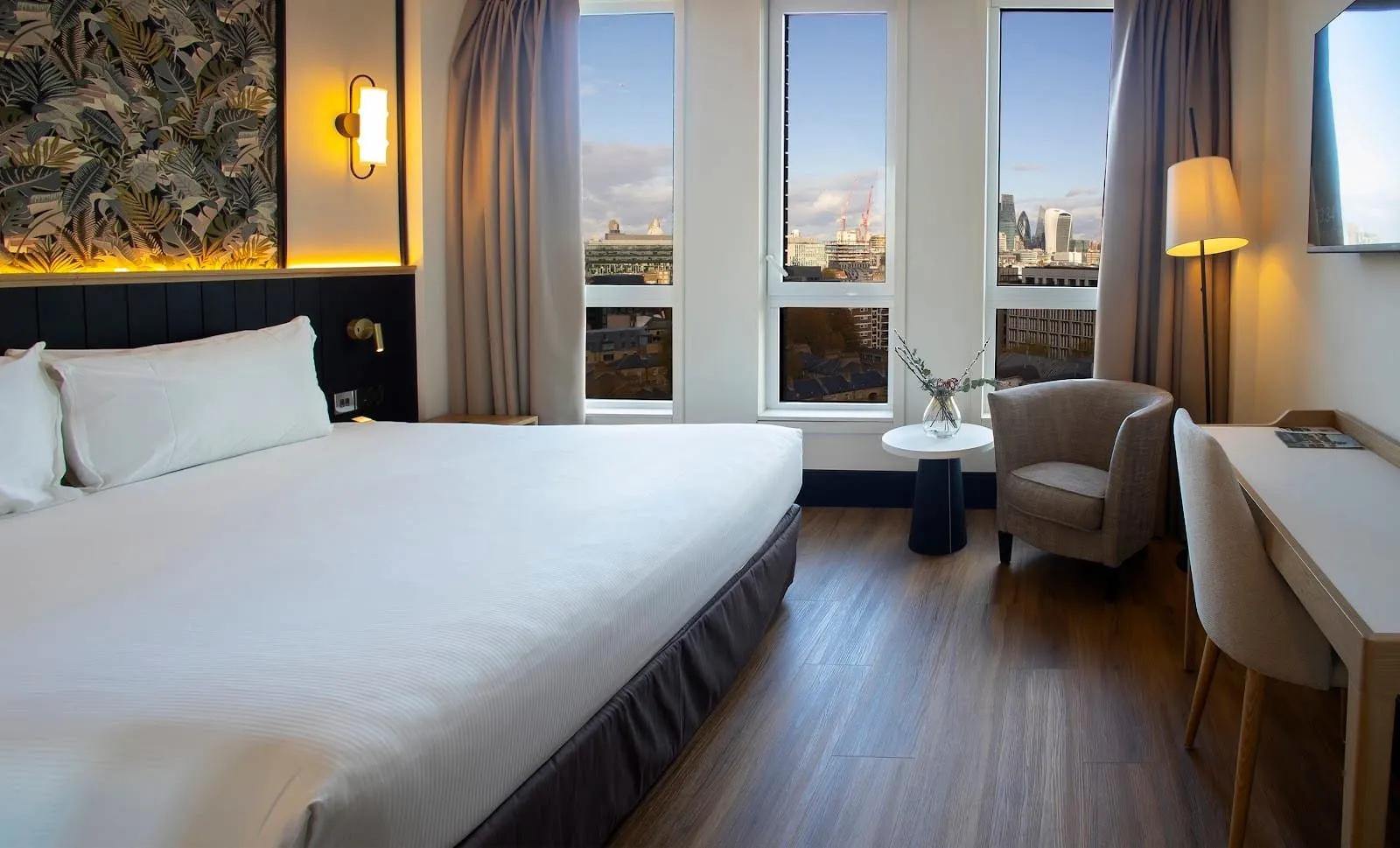 A double room at H10 London Waterloo with a jungle-print panel above the bed, three tall windows onto the City skyline with the Gherkin and the Walkie-Talkie, and a tub armchair