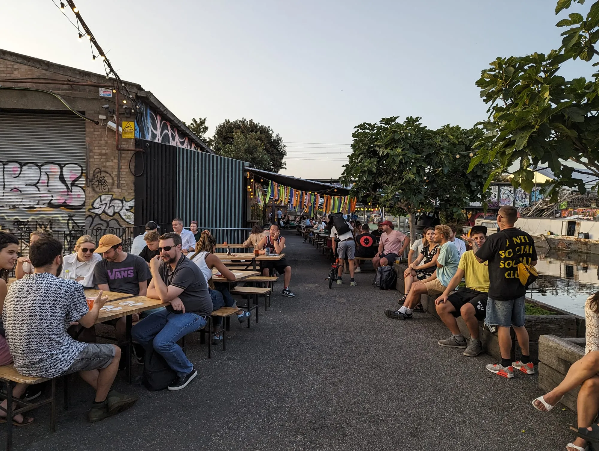People drinking at long canalside tables outside a warehouse bar at Hackney Wick