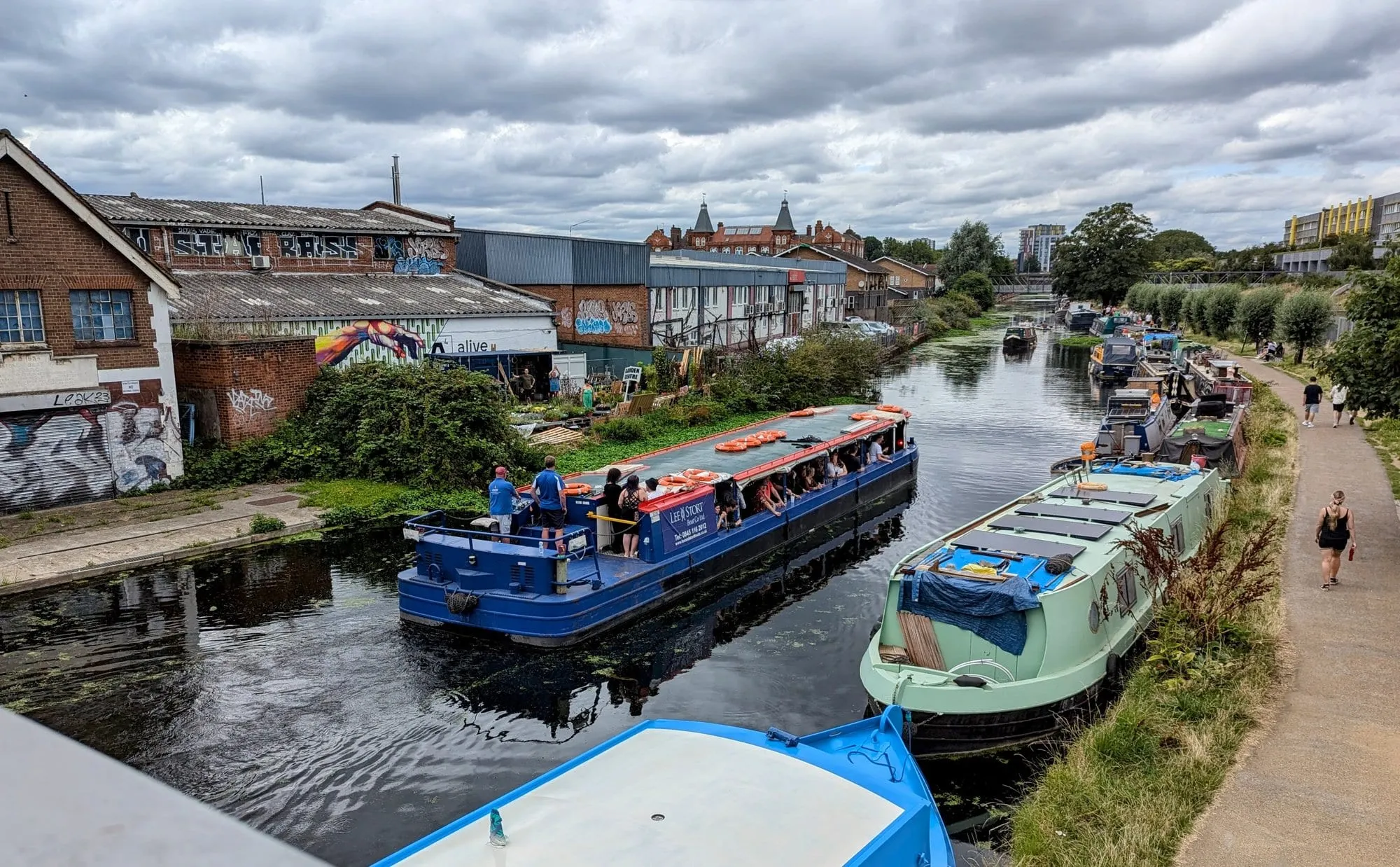 A trip boat crowded with passengers passing moored narrowboats on the Lea Navigation at Hackney Wick, graffiti-covered warehouses on the bank behind