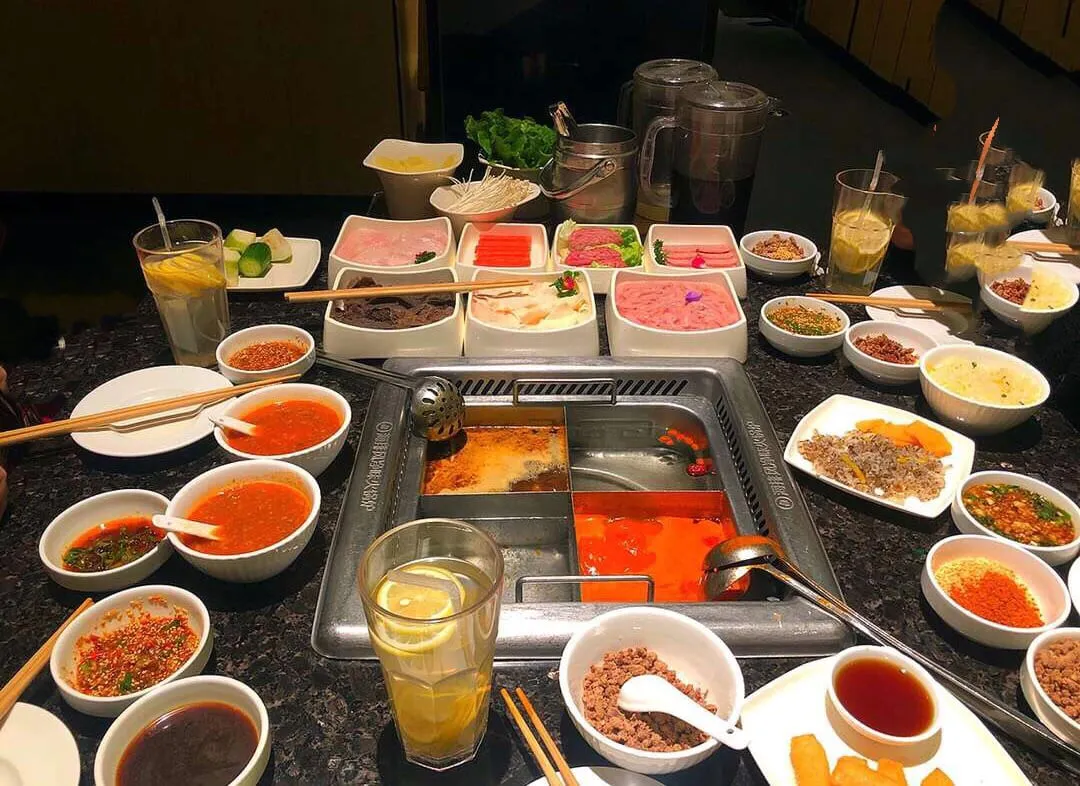 A Haidilao table: a divided rectangular pot of mala and clear broth, raw plates of fish, beef and prawn paste, and a ring of dipping sauces and dry seasonings from the counter