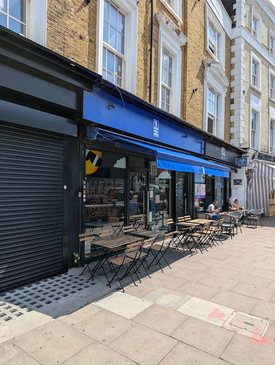 Half Cut's shopfront, with a blue fascia and awning, the name in orange neon in the window, and wooden folding tables and chairs on the pavement