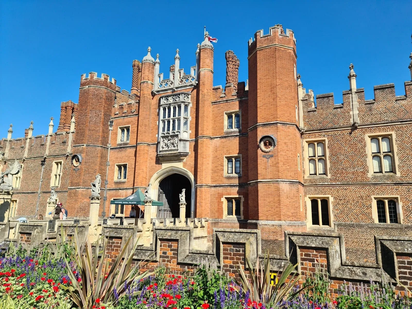 The red-brick Great Gatehouse on the west front of Hampton Court Palace, with a Union flag on its turret and flower beds in front of the moat wall