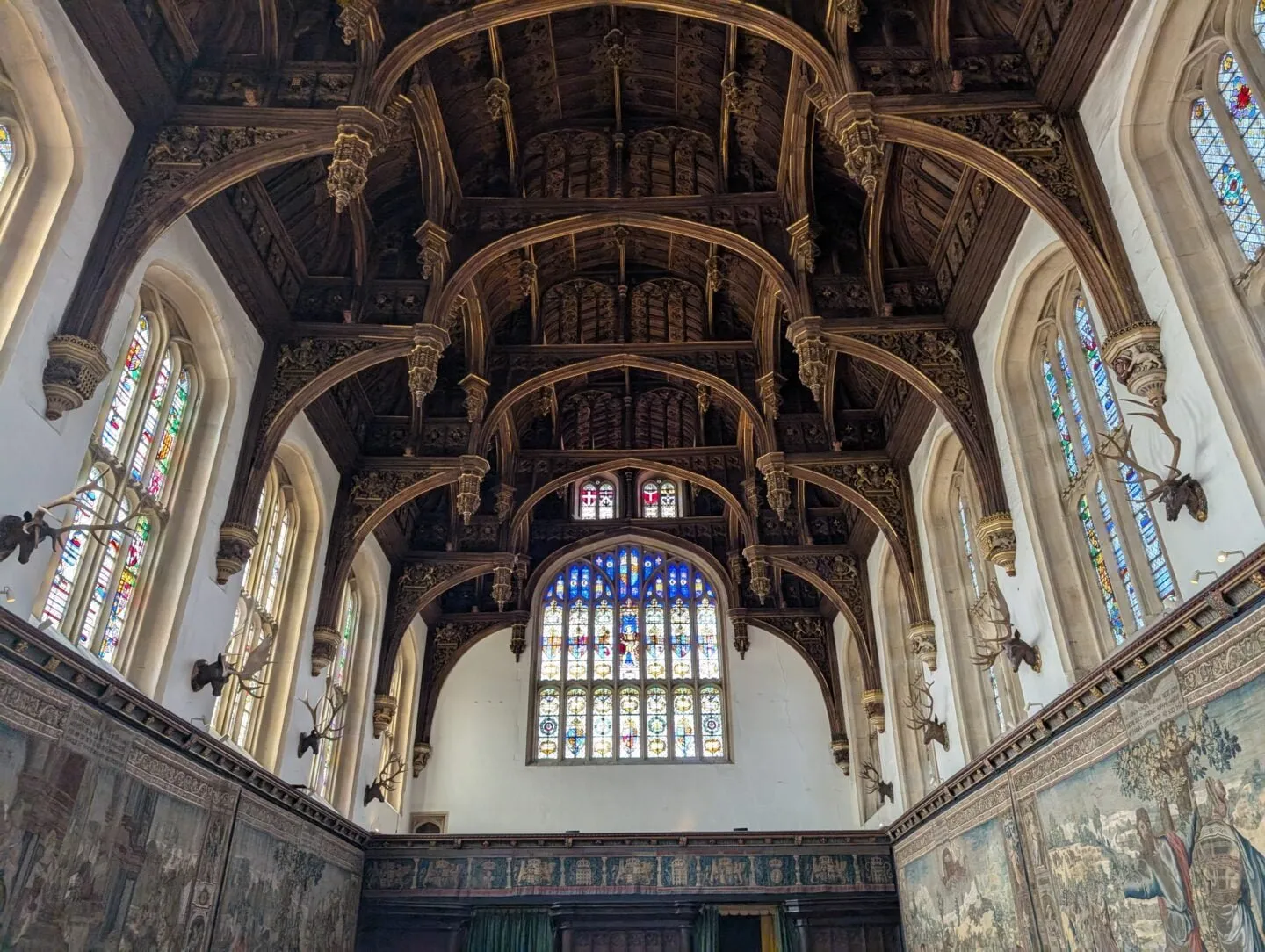 Looking up at the carved wooden hammerbeam roof and stained-glass windows of the Great Hall at Hampton Court Palace, with tapestries and antlers on the walls below