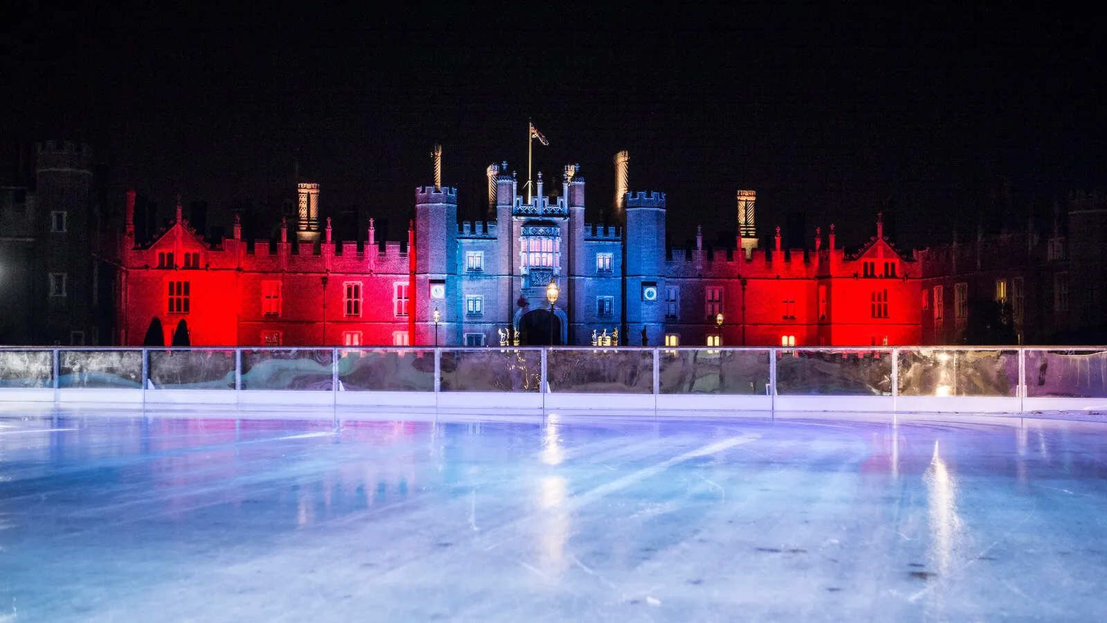 The Hampton Court Palace ice rink at night, empty ice in front of the Tudor West Front floodlit red either side of the blue-lit Great Gatehouse