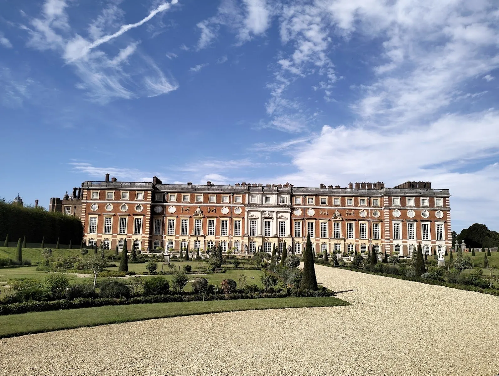Christopher Wren's red-brick and stone range at Hampton Court Palace, seen across a formal garden of clipped yew cones and a gravel path