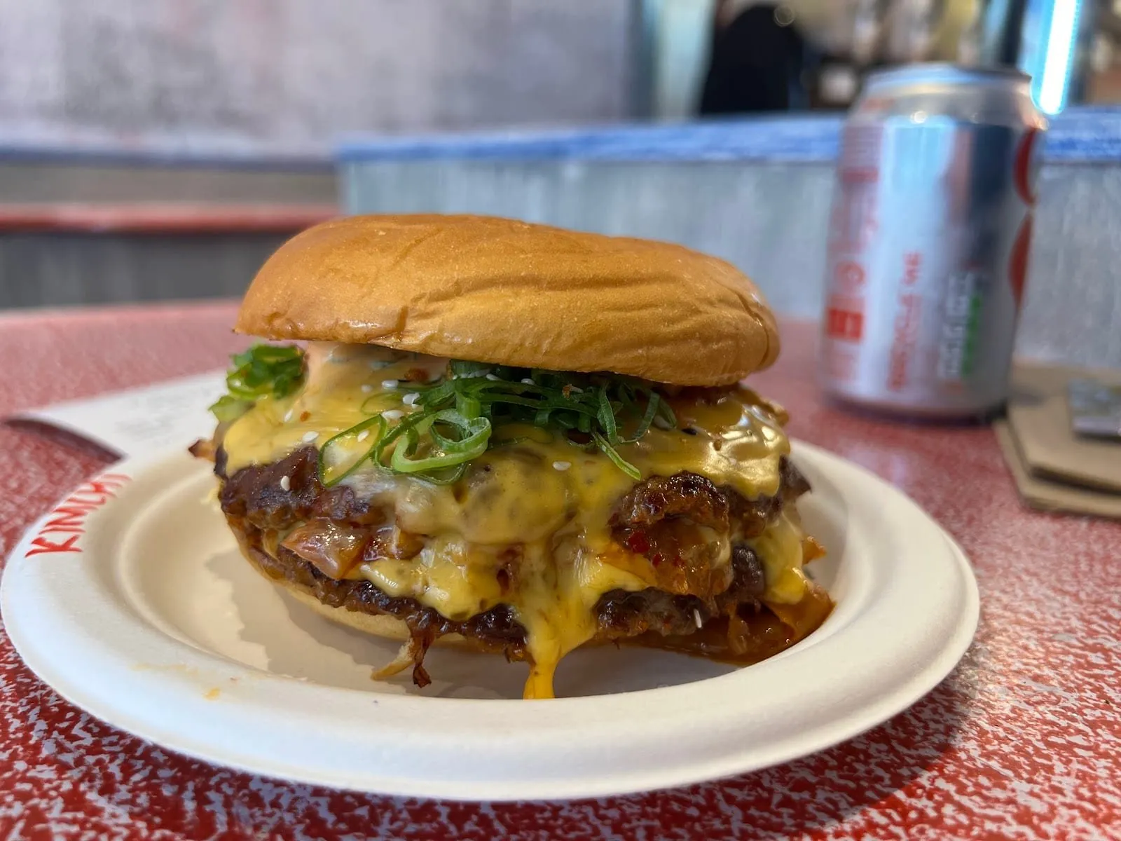 A Hanbaagaasuuteeki double smash burger on a paper plate with "Kimchi" written on the rim in red: two patties under melted cheese, kimchi, sesame seeds and a pile of sliced spring onion in a soft bun, on a red speckled table