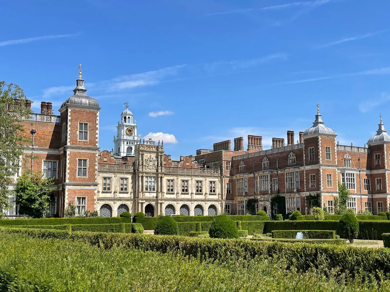 Hatfield House, a red-brick Jacobean mansion with a central white clock tower, seen across its clipped garden hedges