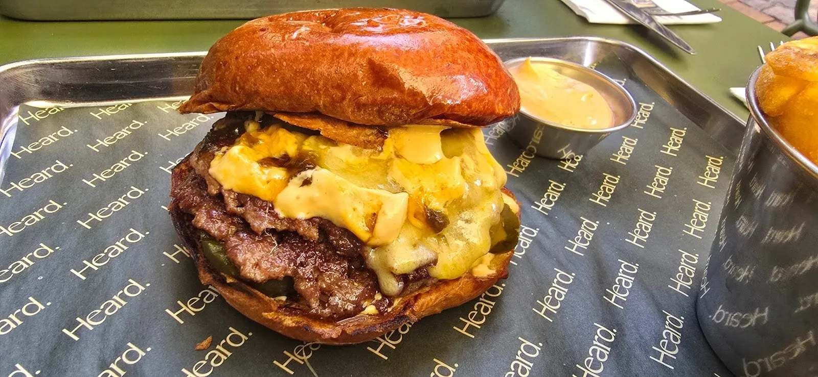 A Heard burger on a metal tray lined with paper printed with the Heard name: two beef patties under a thick layer of melted cheese and pickles in a dark glossy toasted bun, with a pot of sauce and a cup of chips beside it