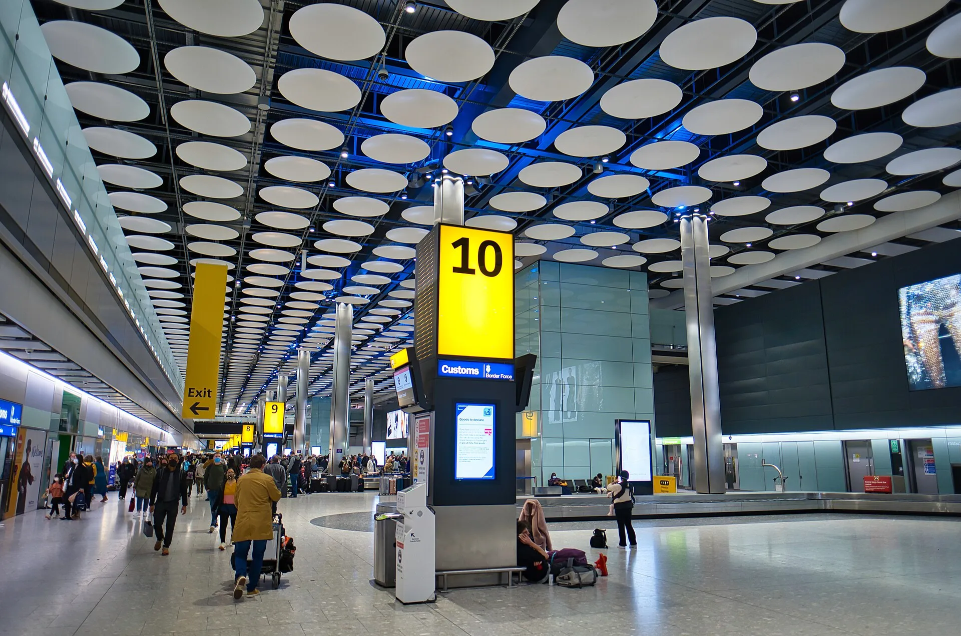 A Heathrow arrivals baggage reclaim hall, carousel 10 under a ceiling of white discs, with the Customs and Border Force channel ahead and passengers walking out towards the exit