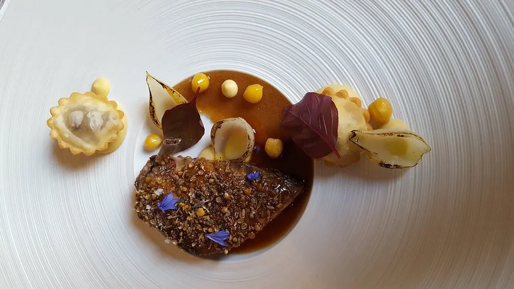 A seared, seed-crusted piece of foie gras in a dark jus, with a small ravioli, charred onion petals, purée dots and a blue cornflower on a ridged white plate