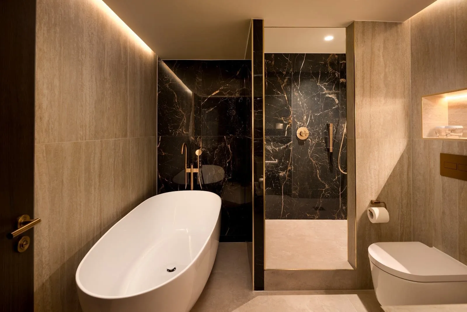 A bathroom at Hilton London Bankside with a freestanding bath, a walk-in shower lined in black marble, brass fittings and travertine-effect walls