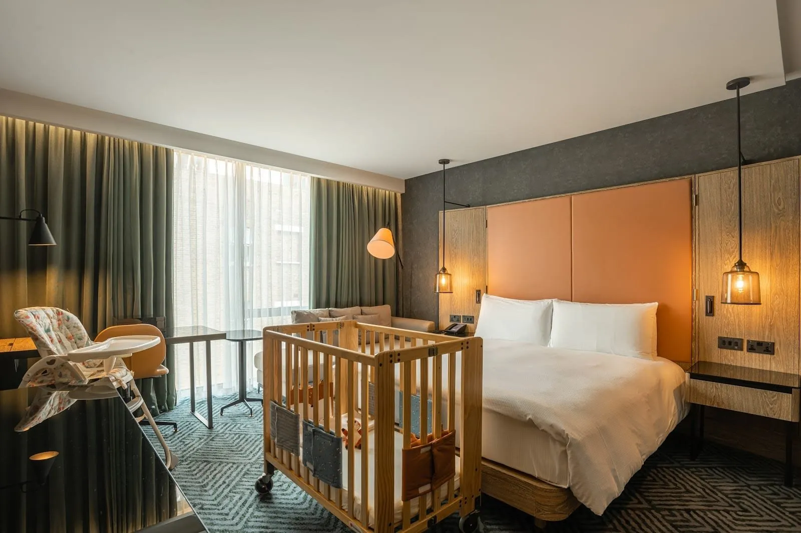 A room at Hilton London Bankside set up for a family, with a wooden cot and a high chair beside a double bed with a tan leather headboard, and a sofa by a full-height window