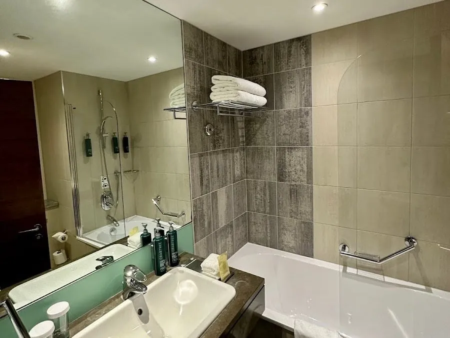 A bathroom at Hilton London Canary Wharf with a bath and shower behind a glass screen, grey wood-effect tiles, a grab rail, and a basin set in a dark counter