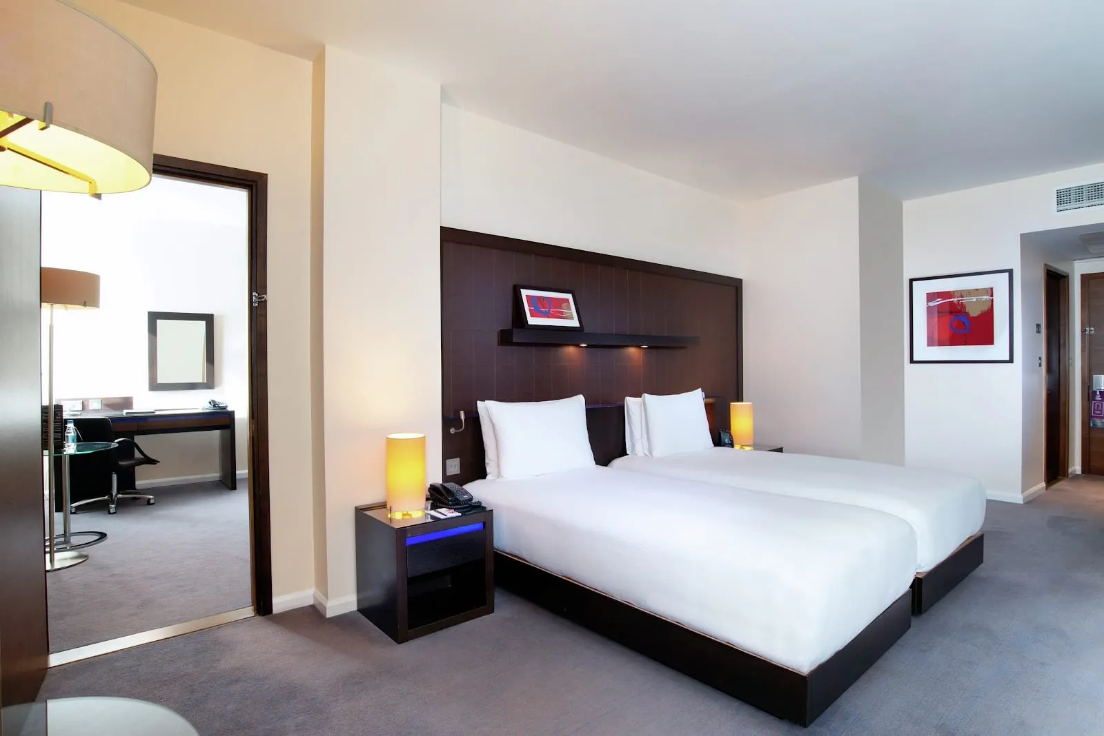 A room at Hilton London Canary Wharf with twin beds against a dark wood headboard wall, yellow bedside lamps, and a doorway through to a separate study with a desk and office chair