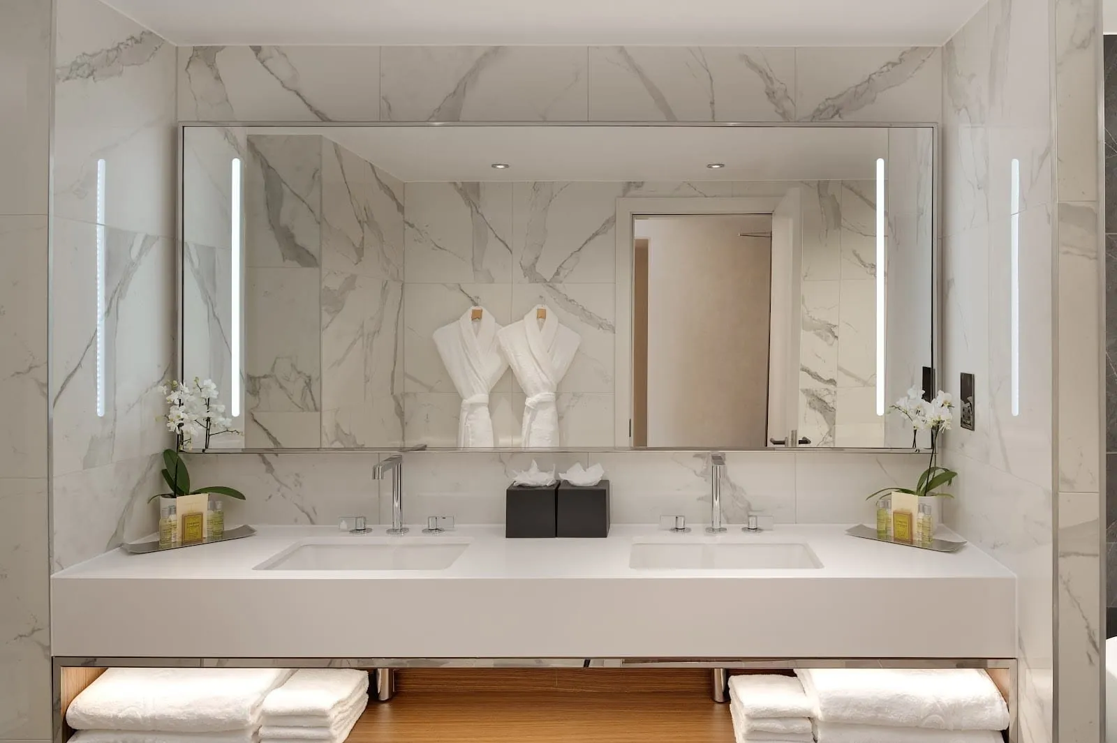 A white marble bathroom at Hilton London Metropole with twin basins under a wide lit mirror, two bathrobes reflected on the wall and towels stacked beneath the counter